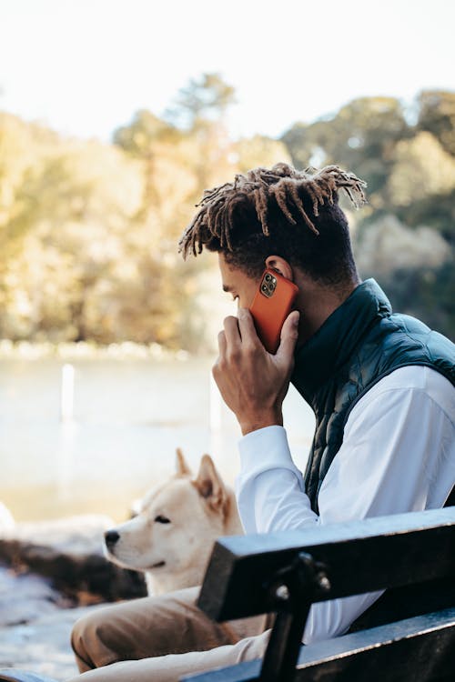 Side view of unrecognizable young ethnic guy with dreadlocks in stylish outfit having phone conversation while resting in park near pond with adorable Akita dog