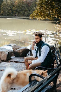 From above of focused African American male speaking on smartphone while sitting on bench near calm lake with loyal brown dog on leash