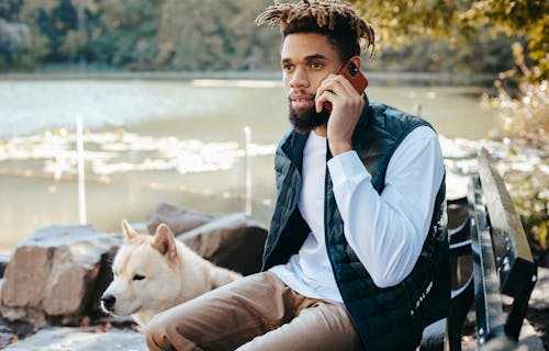 Free Serious bearded African American male having phone call while sitting on bench near dog at calm lake in park on sunny weather Stock Photo