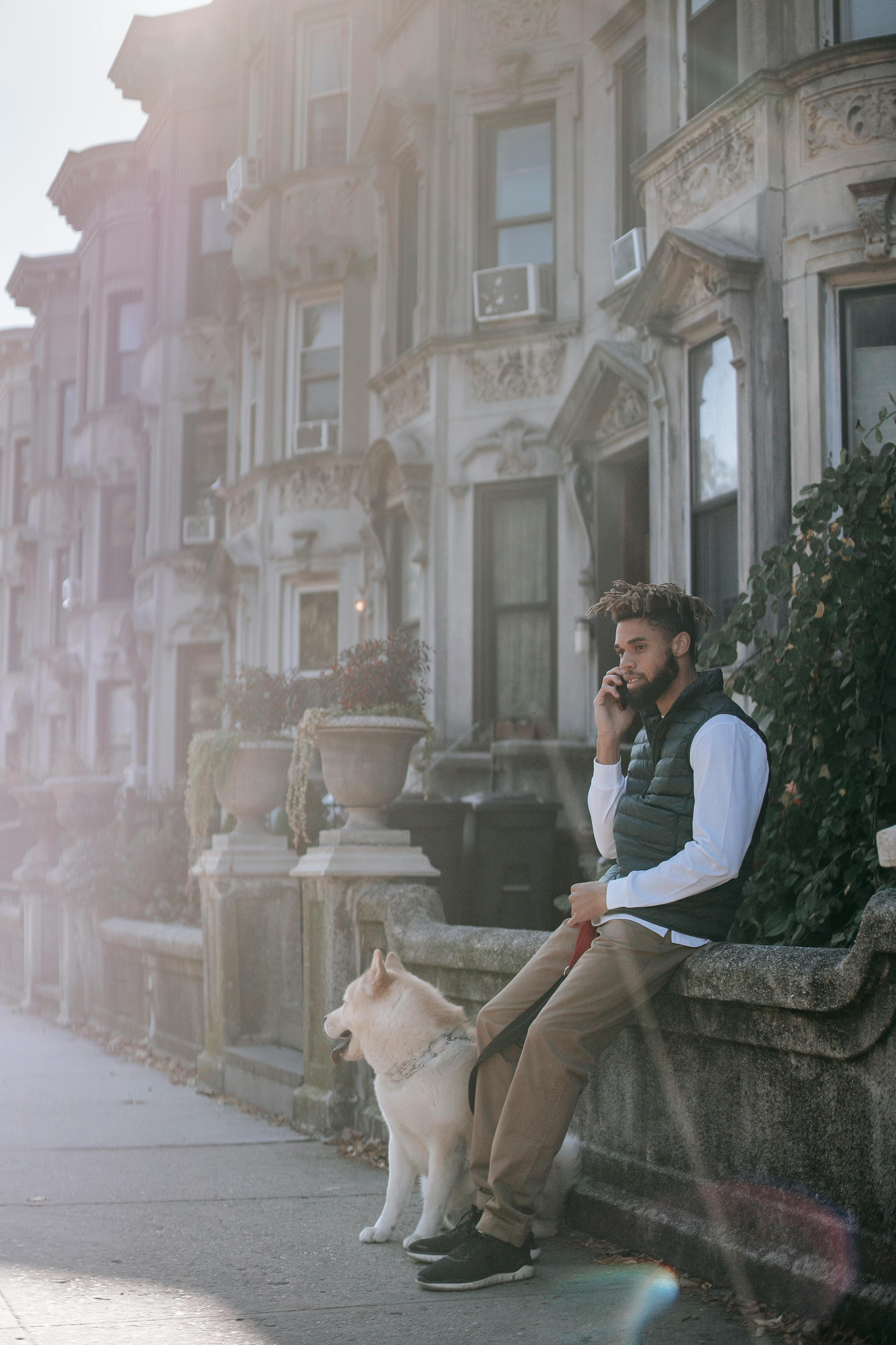 black man having phone conversation near dog while sitting near building