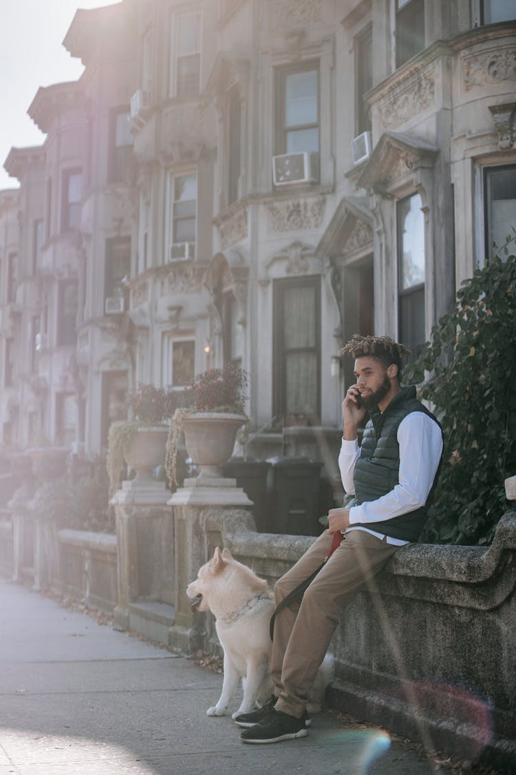 Black Man Having Phone Conversation Near Dog While Sitting Near Building