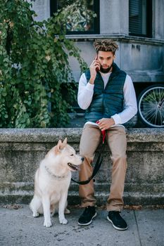 在城市里，一名男子坐在一只秋田犬旁边，正用智能手机通话
