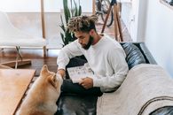 Side view of bearded African American male reading book while sitting on couch near obedient dog in room with table