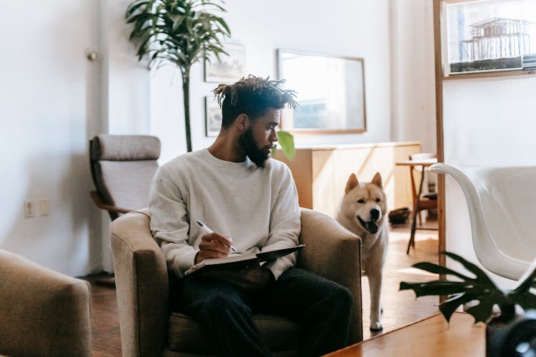 Serious Black Man Writing In Notebook In Room With Dog