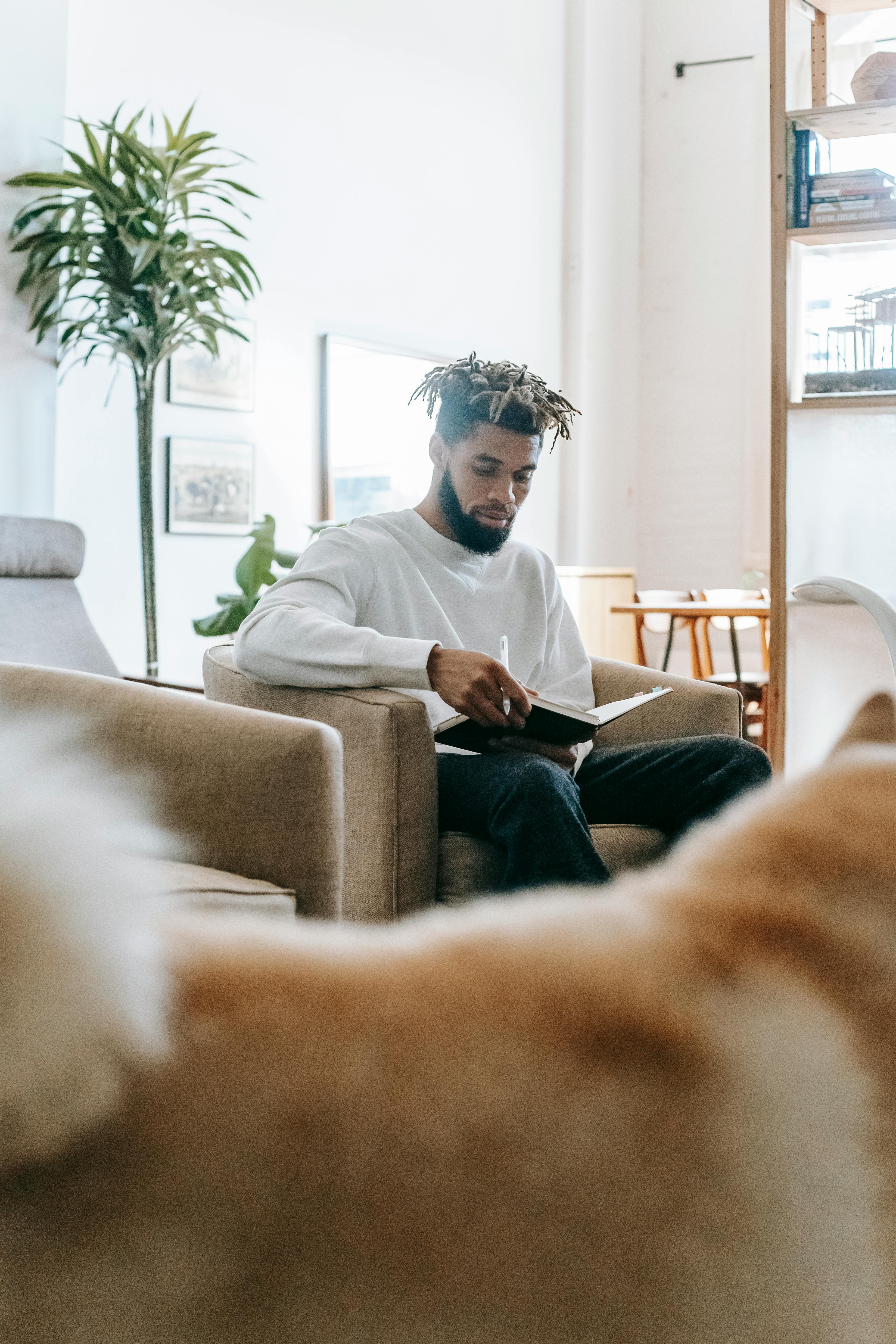 Focused bearded African American male with dreadlocks sitting in armchair with notepad and pen in living room with shaggy dog