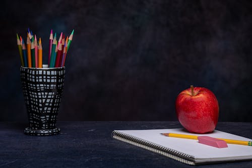 Set of multicolored pencils in cup holder placed on table with eraser on copybook and ripe red apple on black background