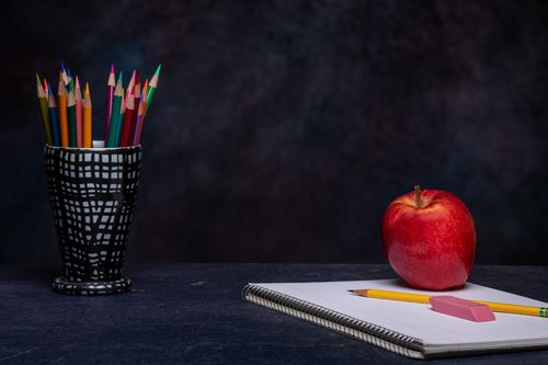 Free Set of multicolored pencils in cup holder placed on table with eraser on copybook and ripe red apple on black background Stock Photo