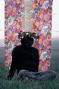 Full body anonymous person in dark casual wear and black incognito mask wearing wreath on head and sitting on grassy meadow against building wall with floral ornament