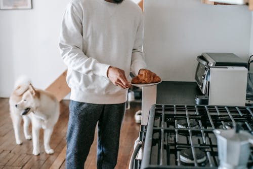 Faceless man with croissant in hand standing in kitchen with cute dog