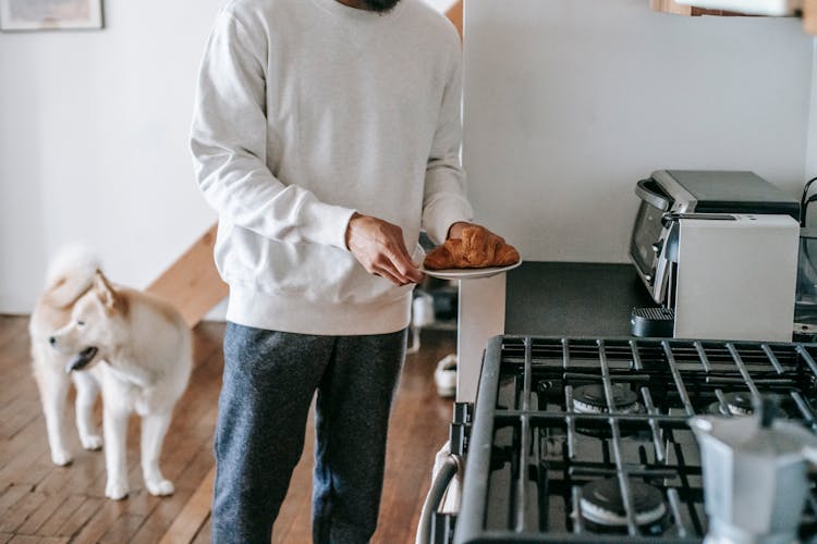 Faceless Man With Croissant In Hand Standing In Kitchen With Cute Dog