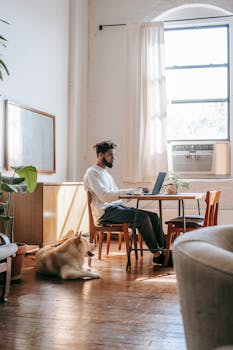 A man working remotely on a laptop in a cozy room with a dog lying nearby.