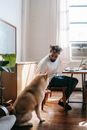 Young ethnic guy playing with pet while using laptop at home