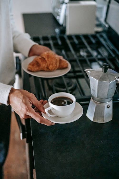 Free From above of crop unrecognizable person in casual clothes holding plate with fresh croissant and cup of coffee brewed in moka pot before breakfast in kitchen Stock Photo