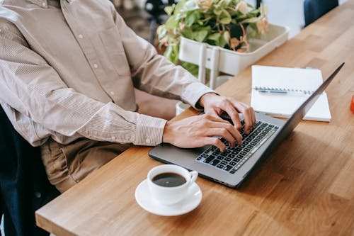 High angle of crop anonymous male freelancer in casual clothes sitting at table and using netbook while drinking cup of coffee and notebook