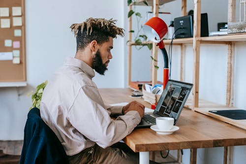 Side view of African American male freelancer in casual outfit sitting at wooden table and working on laptop near cup of coffee and lamp near shelves in light room