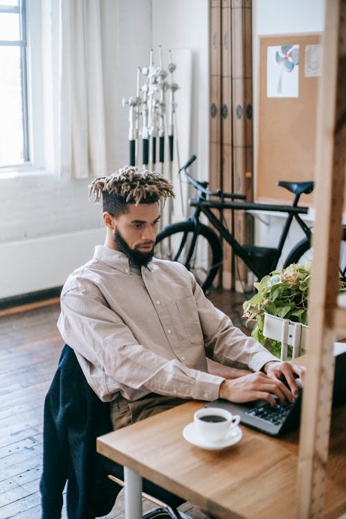 Free Serious African American male freelancer in casual clothes sitting at wooden table while working on netbook with cup of coffee and bicycle in workplace Stock Photo