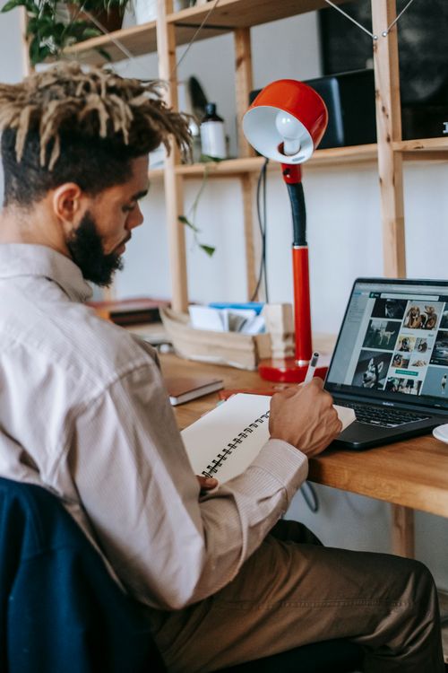 Free Side view of African American man freelancer in casual clothes working on laptop while sitting at wooden table and making notes in notebook near lamp in room Stock Photo