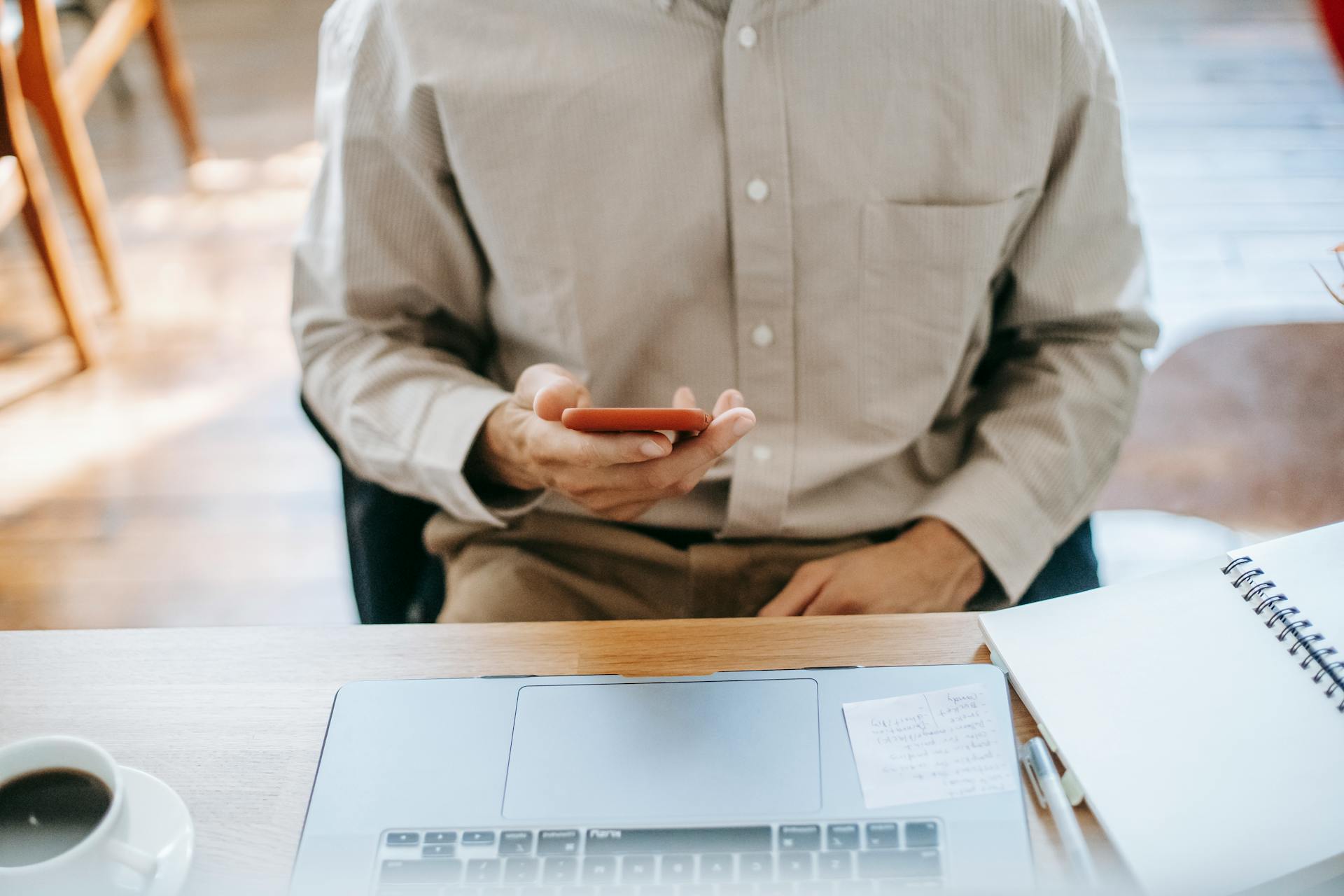 Crop unrecognizable man in casual clothes working remotely on computer while sitting at wooden table and checking notifications on phone near notebook and cup of coffee in light room