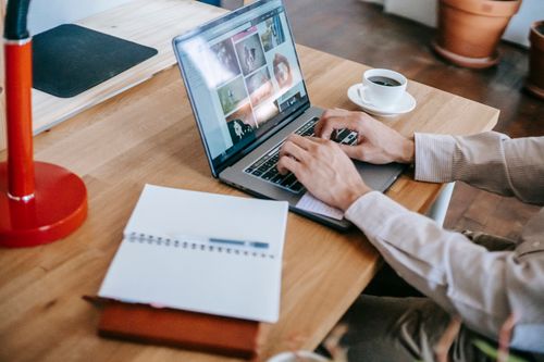 Free High angle of crop faceless male remote worker in casual clothes sitting at wooden table while using laptop near notepad and drinking coffee in light room Stock Photo