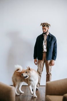 一位穿着休闲服装的非裔美国男子和一只秋田犬站在明亮的房间里，旁边有一把扶手椅，墙壁是白色的