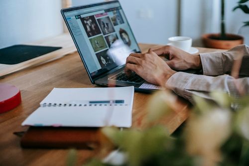 Free Crop unrecognizable guy in casual outfit working remotely on computer while sitting at wooden table near notebook with cup of coffee in light room Stock Photo