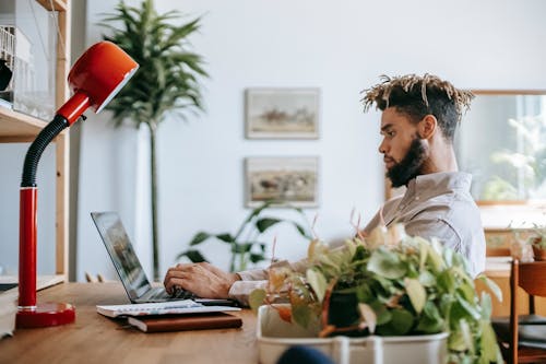 Free Side view of concentrated black guy in casual outfit sitting at wooden table while working remotely on computer with notepad and lamp in workspace Stock Photo
