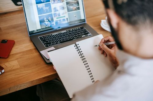 Free High angle back view of faceless male in casual outfit working remotely on netbook while sitting at wooden table and taking notes in notepad with pen near phone in room Stock Photo