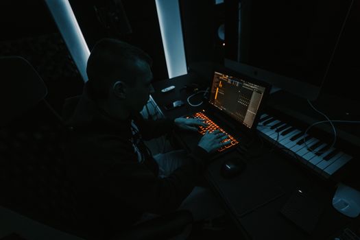 Music producer focused on composing tracks in a dimly lit studio with laptop and keyboard.