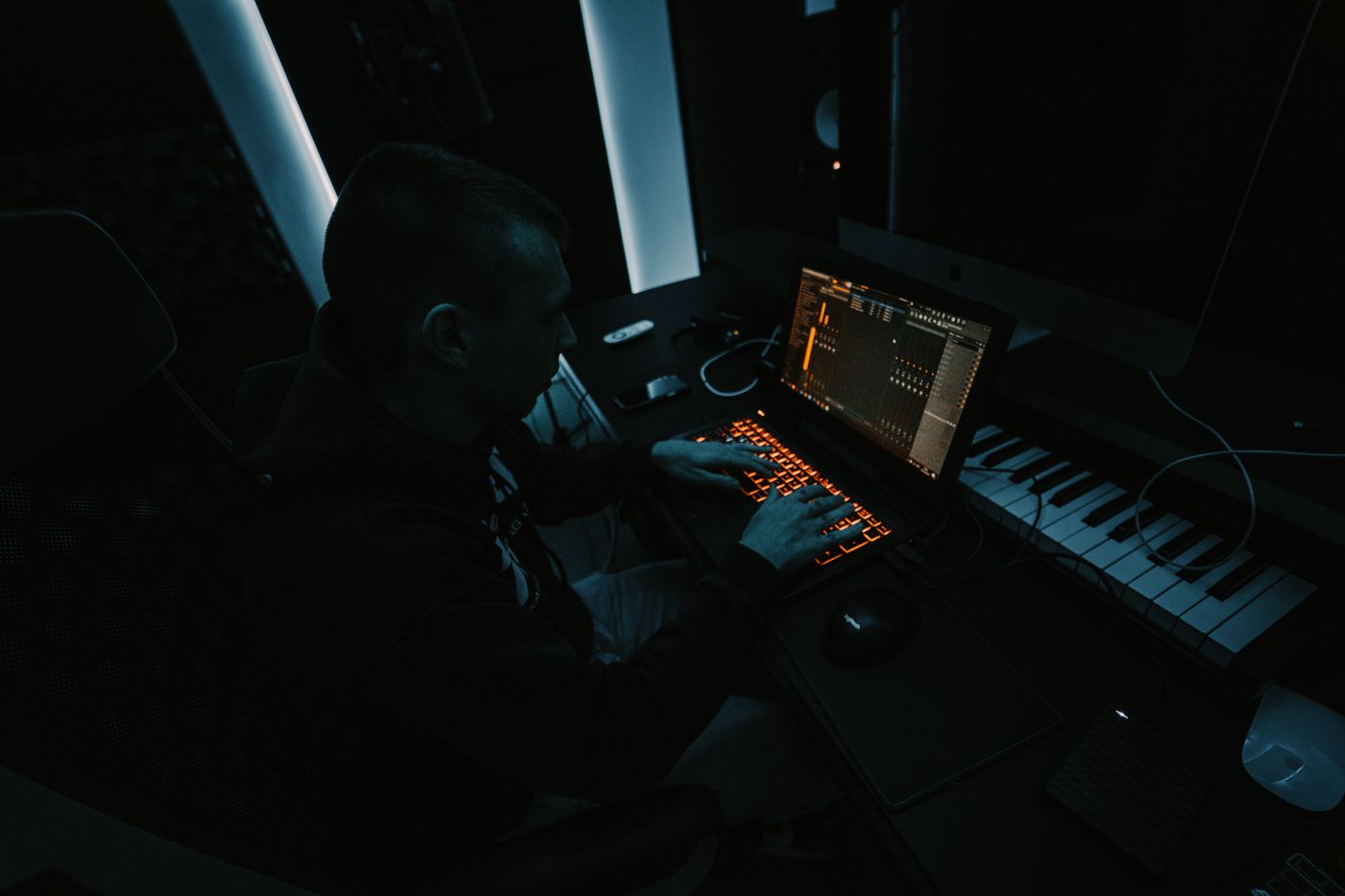 A silhouetted producer typing on a laptop beside a MIDI keyboard in a dark room