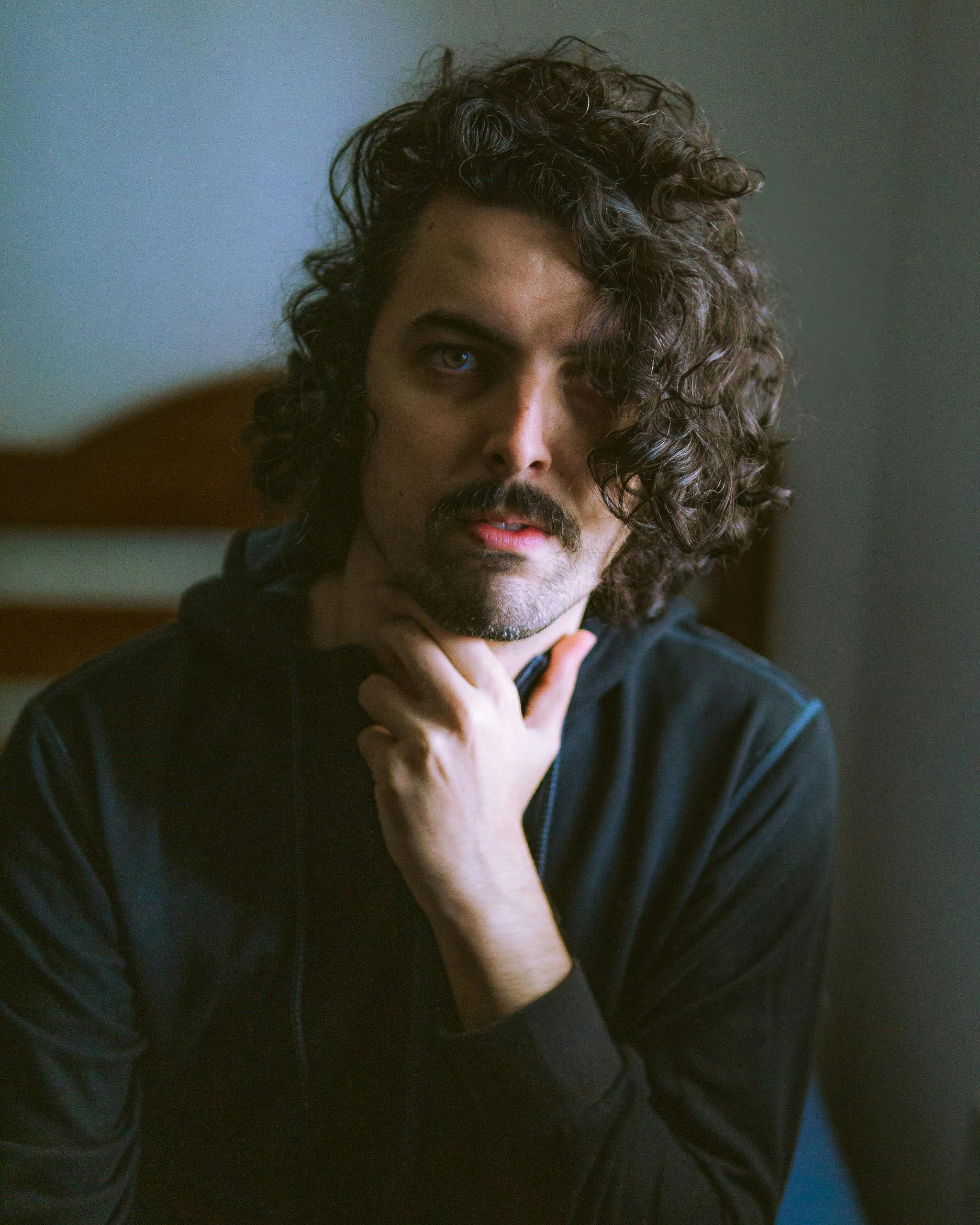 Moody portrait of a stylish man with wavy hair and beard indoors, posing thoughtfully.