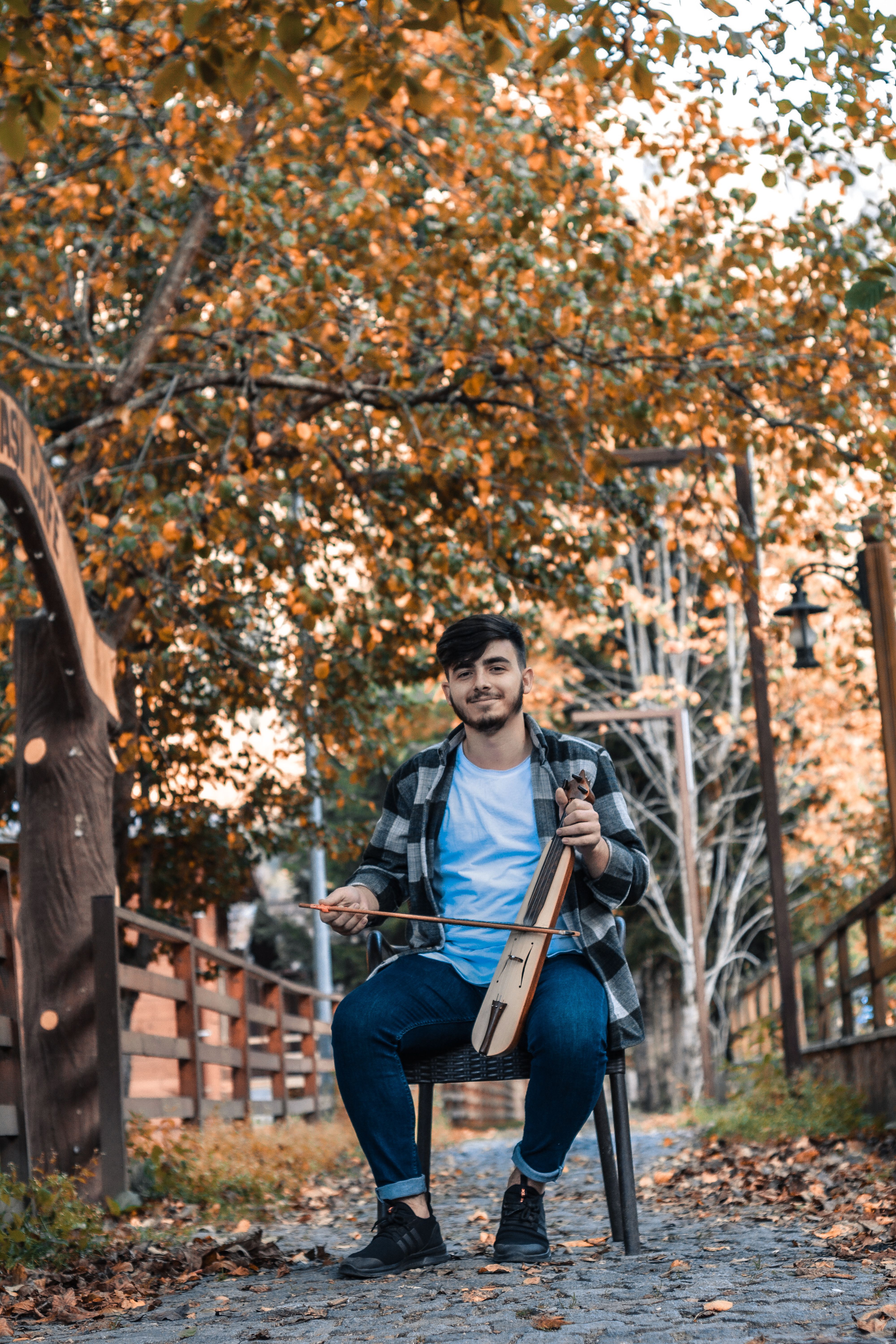 A smiling young man playing a kemence in a park with autumn colors.