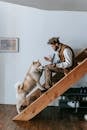 Side view full body of African American guy in casual clothes sitting on stairway in house using laptop with Akita Inu dog disturbing