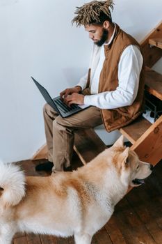 一名男子轻松地浏览着笔记本电脑，旁边的木楼梯上站着一只秋田犬