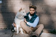 一个穿着休闲服的黑人男子，在晴天里和秋田犬一起坐在砖墙附近的地上