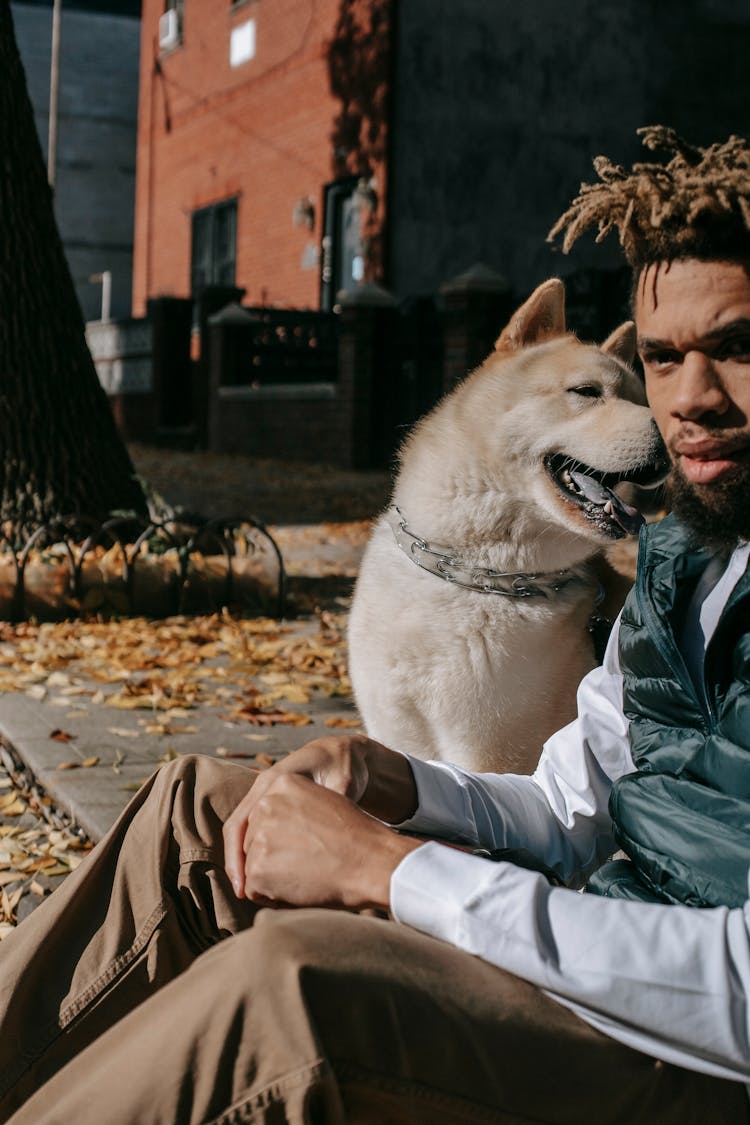 Serious Black Man Sitting On Asphalt With Dog On Street