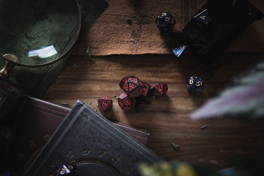 A dark, moody still life scene featuring red and blue gaming dice, books, and a magnifier on a wooden surface.