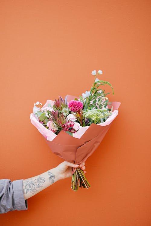 Unrecognizable tattooed person with colorful various flowers with long stems wrapped in paper in hand on orange background on street