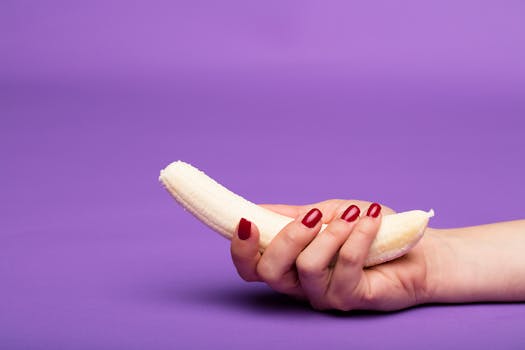 A hand with manicured nails holds a peeled banana against a vibrant purple background in a studio setup.