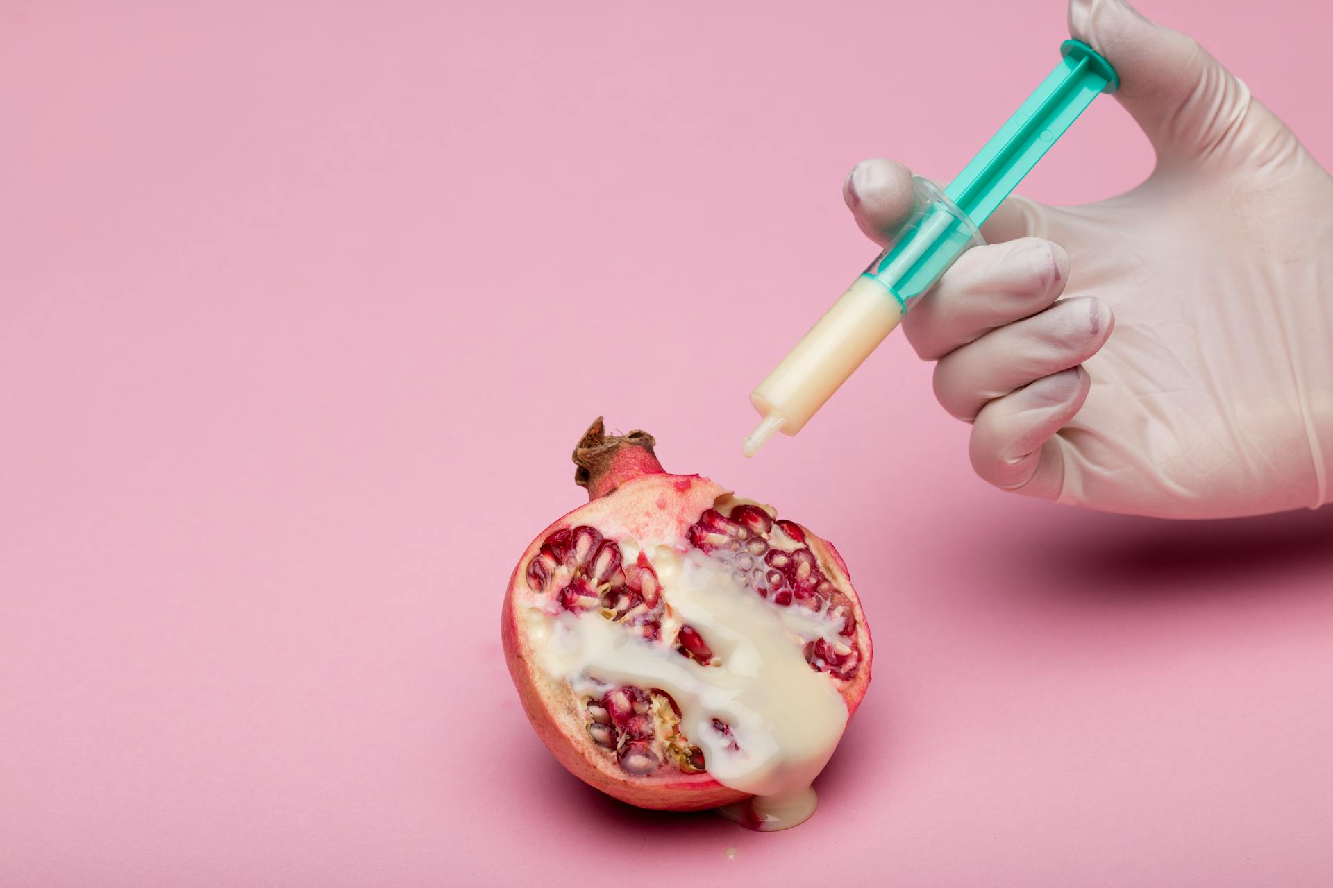 A pomegranate injected with liquid using a syringe held by a gloved hand, set against a pink background.
