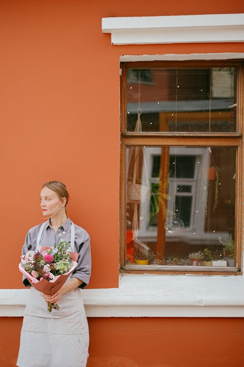 Calm female in apron standing with bouquet of flowers in wrapping paper standing at orange house with window on street