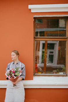 一位平静的女性系着围裙，手捧一束鲜花，站在街上那栋有窗户的橙色房子里