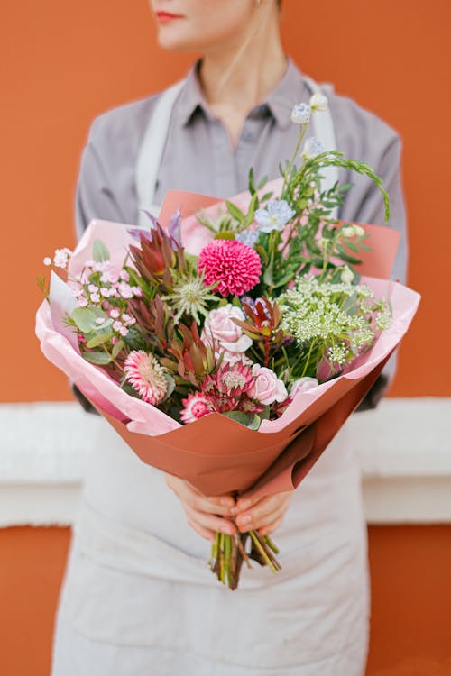Unrecognizable female florist in apron standing with various colorful flowers in wrapping paper in hands near orange wall on street