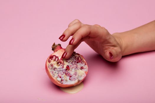 A hand with painted nails delicately touches a pomegranate on a pink background.