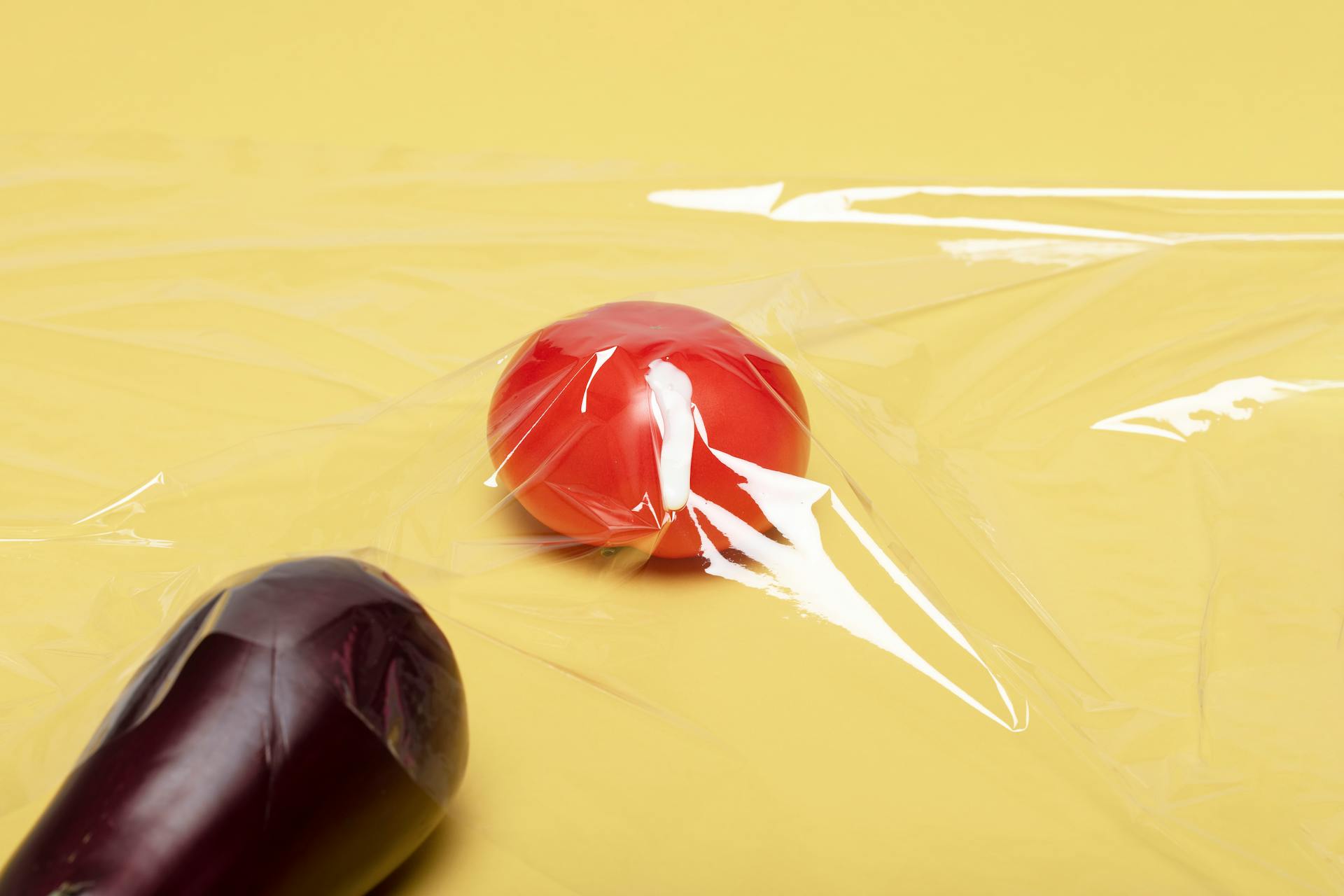 Creative studio shot of a tomato and eggplant on a yellow background, wrapped in plastic film.