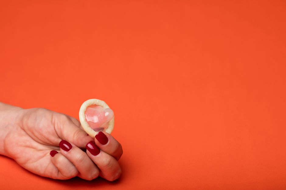 Close-up of a hand with red nails holding a condom on an orange background.