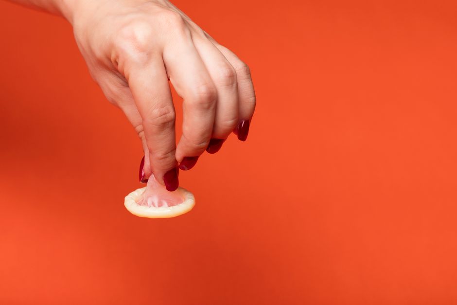 A hand with painted nails holding a condom, set against a vibrant red background.