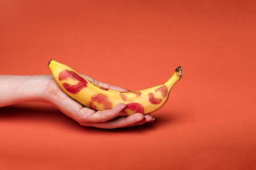 A close-up of a hand holding a banana with lipstick kiss marks against a red background.