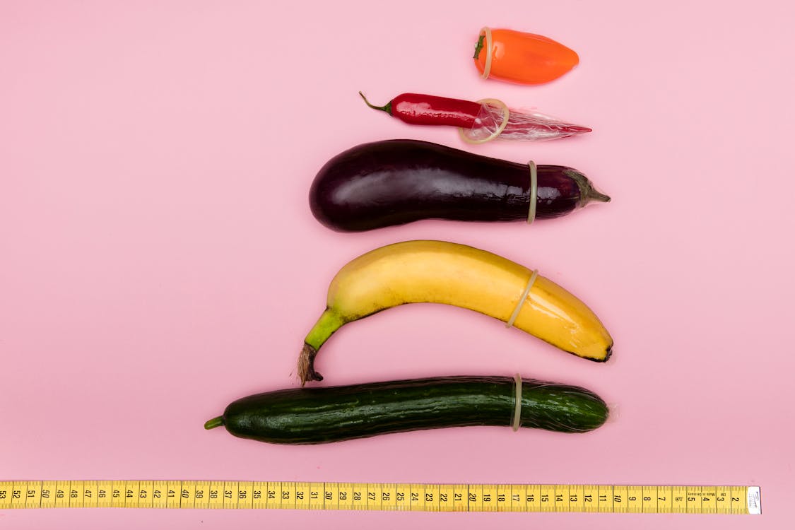A variety of fruits and vegetables, such as a cucumber, banana, eggplant, and chili peppers with a condom on each one. There is a measuring tape below all of the fruits and vegetables.