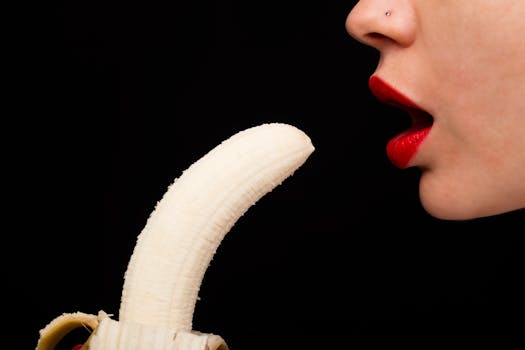 Close-up of a woman with bright red lipstick poised near a peeled banana on a black background.