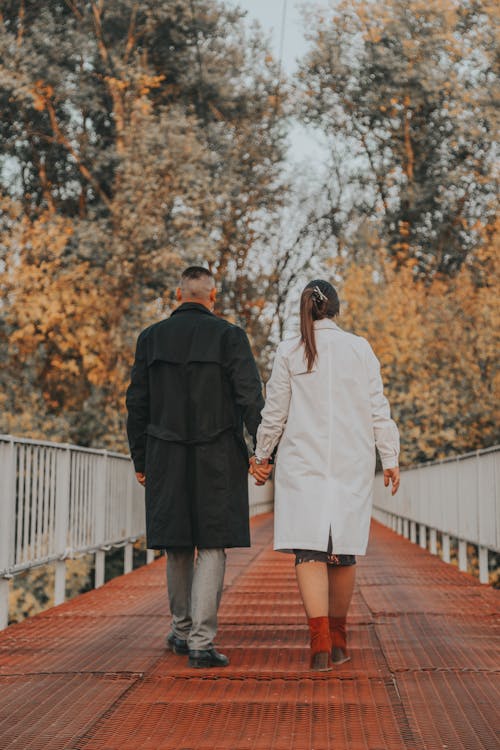 Back view of anonymous man and woman holding hands while walking along bridge in countryside in autumn